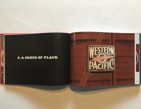 In this colour photograph taken by the railfan designer Ian Logan, a rusting enamel sign screwed to the side of a wooden boxcar carries the name Western Pacific picked out in white serif capitals on a black background with a red feather pointing through a central circle which carries the legend Feather River Route.