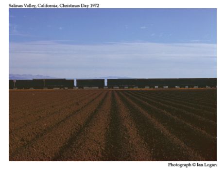 In this colour photograph taken by the railfan designer Ian Logan in the 1970s, a freight train rumbles across asparagus fields in Salinas Valley, California, with ploughed furrows lending perspective to the foreground and blue-grey mountains, hazy clouds and a deep blue sky forming the backdrop.
