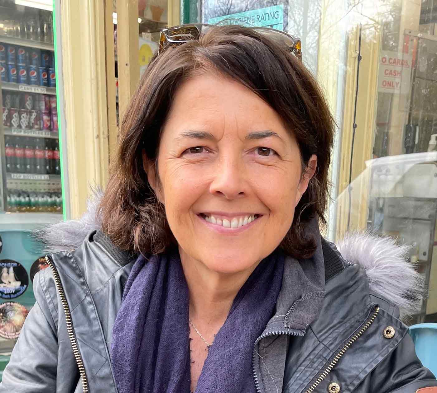 In this colour photograph Cath Harries looks at the camera and smiles as she pauses for a photograph while out door hunting, wearing a dark blue leather jacket and a scarf.