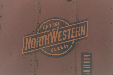 The ball-and-bar logo of the Chicago & North Western Railway is painted in ochre on the side of a brown boxcar, punctuated by rows of rivets and accompanied by an early form of bar code.