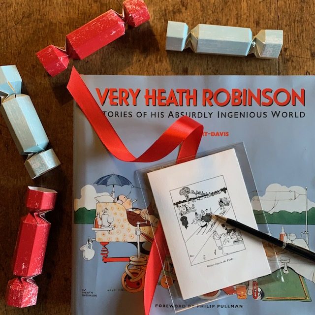 Colour photograph of a Very Heath Robinson book and a pack of six Winter Joys Christmas cards, surrounded by blue and red Christmas crackers.
