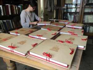 In this colour photograph taken at Sheldrake Press, the editor Kezia Bayard-White assembles the wooden cases of the Very Heath Robinson De Luxe Edition on a table and secures them with knotted string.